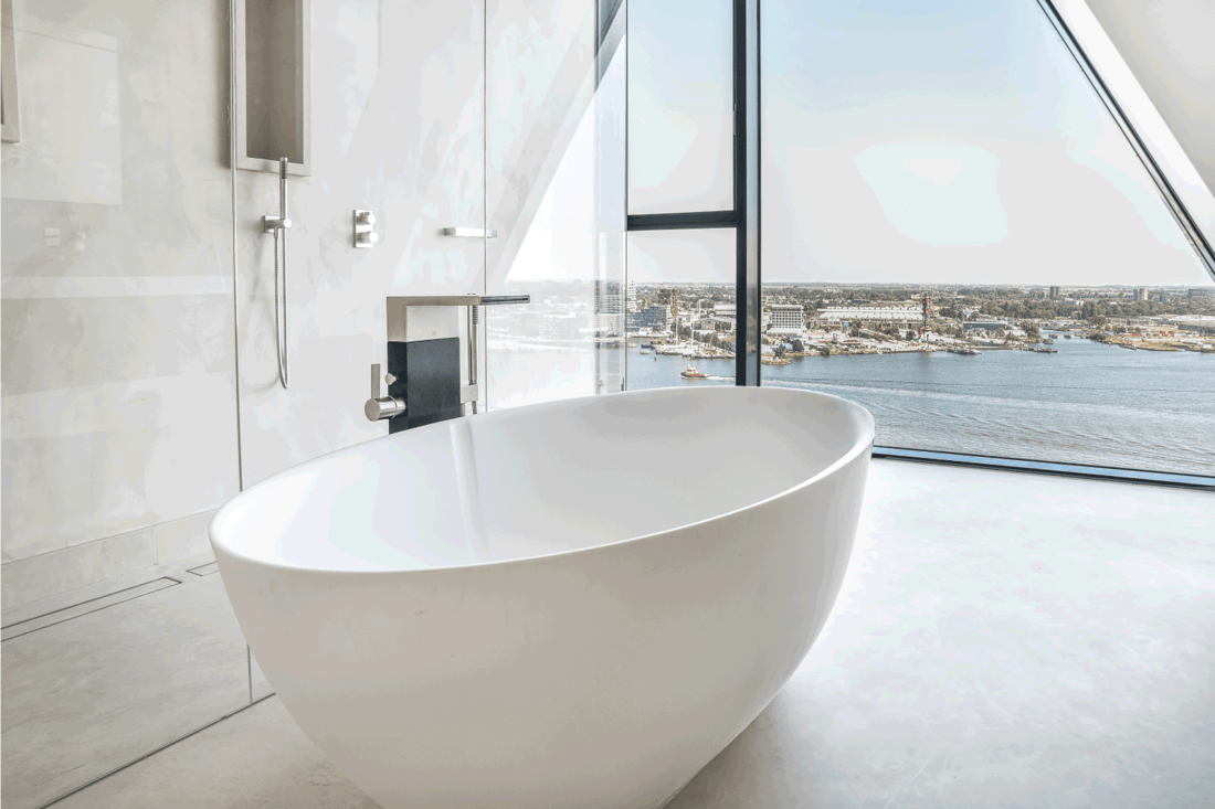 Big white soaking bathtub in spacious bathroom with shower cabin and wall window viewing city, soaking tub