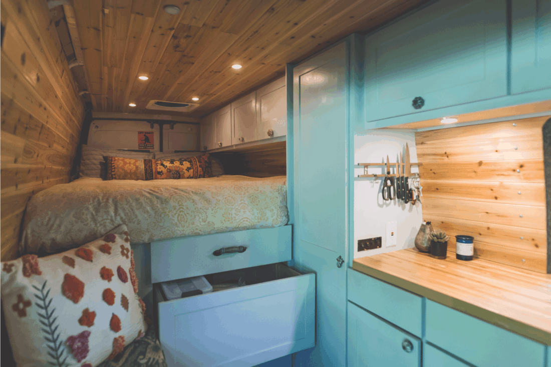 Living space of a young couple who live in a van. The sleeping area and bed are on the left. Underneath are cupboards and storage space.