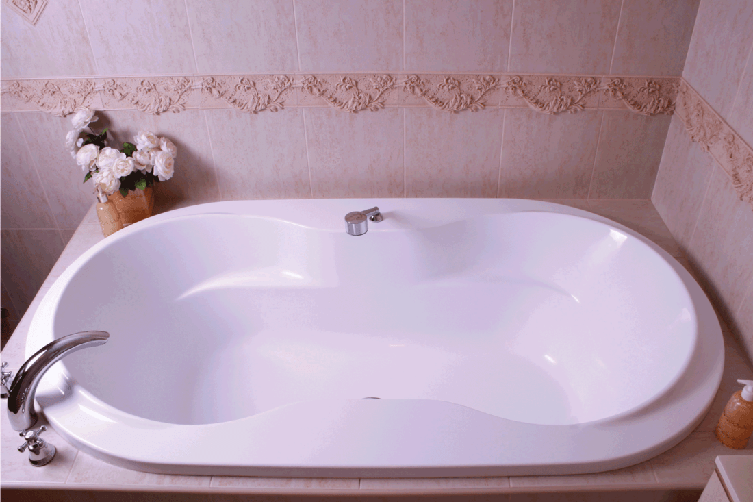 White bath tub with faucet and beige tiles in bathroom. Oval shaped corner bathtub