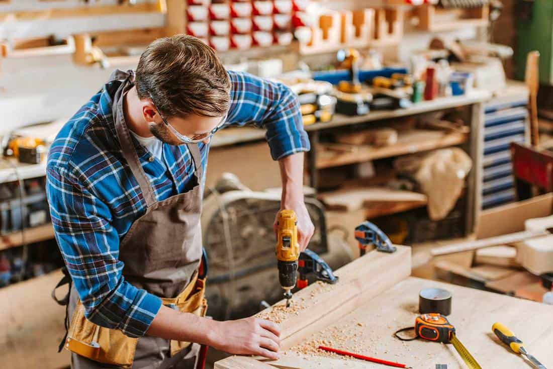 Woodworker in goggles and apron holding hammer drill near wooden planks
