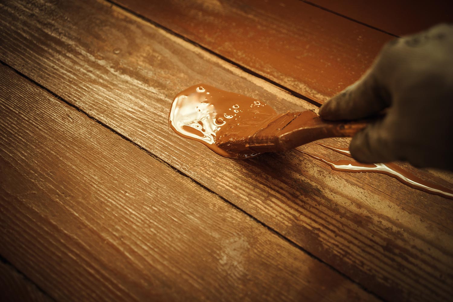A hand in a gray glove with a brush paints the floor with brown oil paint