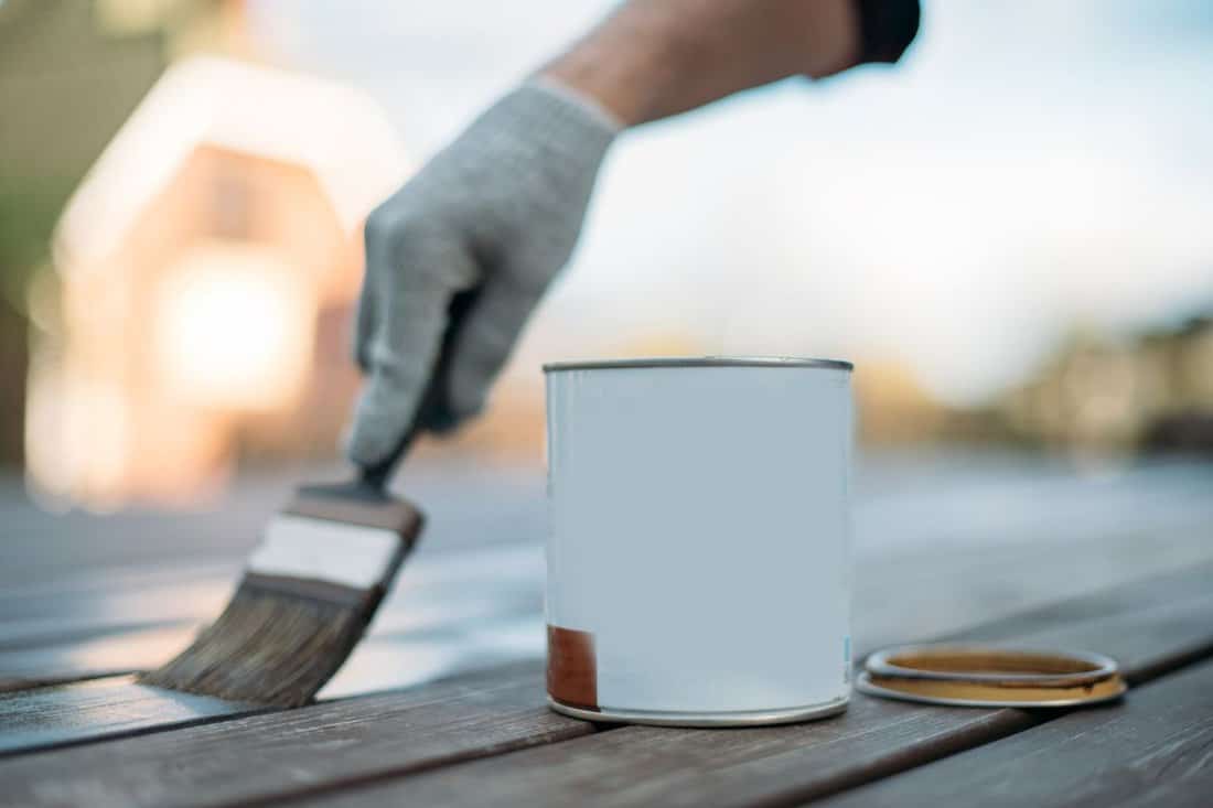 Close-up, an open jar and a man's hand in a work glove with a painting brush paints boards outdoors. A hand applies paint, oil or varnish to the veranda floorboards