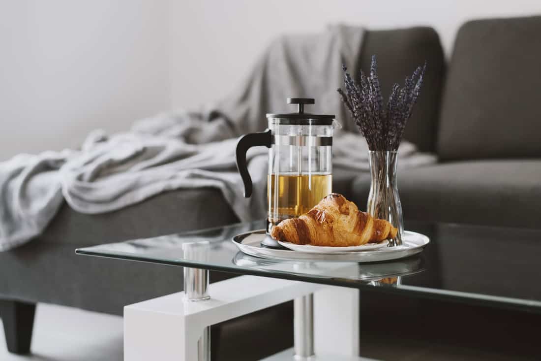 Contemporary interior living room with a glass coffee table, a small plate with croissant, French press, and a vase with lavender
