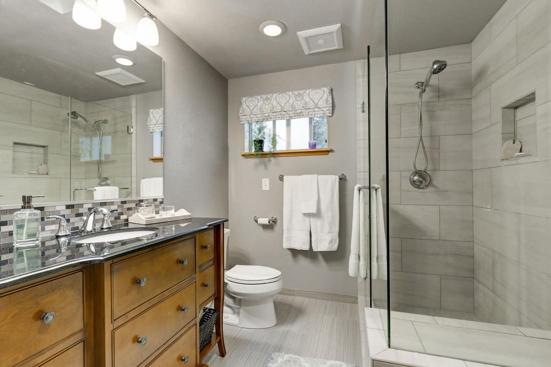 Contemporary interior of a bathroom with light gray colored walls, glass shower wall, and wooden cabinetry and a huge mirror on the vanity, What Color Vanity Goes With Grey Walls?
