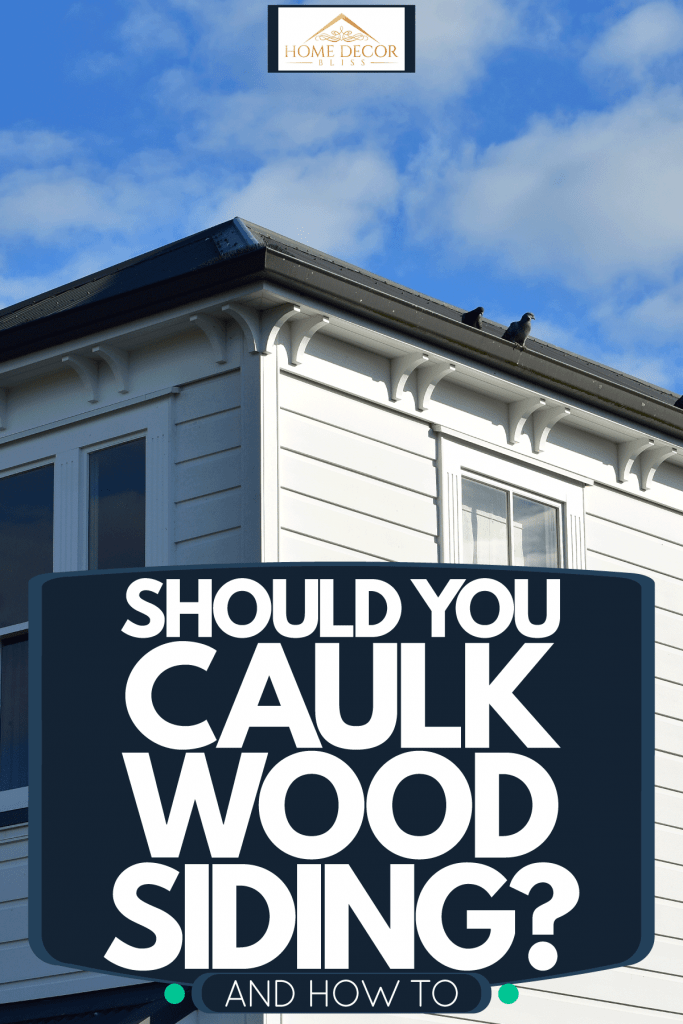 Gray wooden siding of a ranch style house with white painted window frames, Should You Caulk Wood Siding? [And How To]
