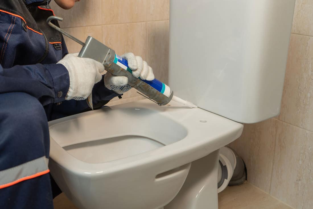 A worker caulking the toilet tank, How To Fix A Cracked Toilet Tank, Bowl, Or Base [A Complete Guide]