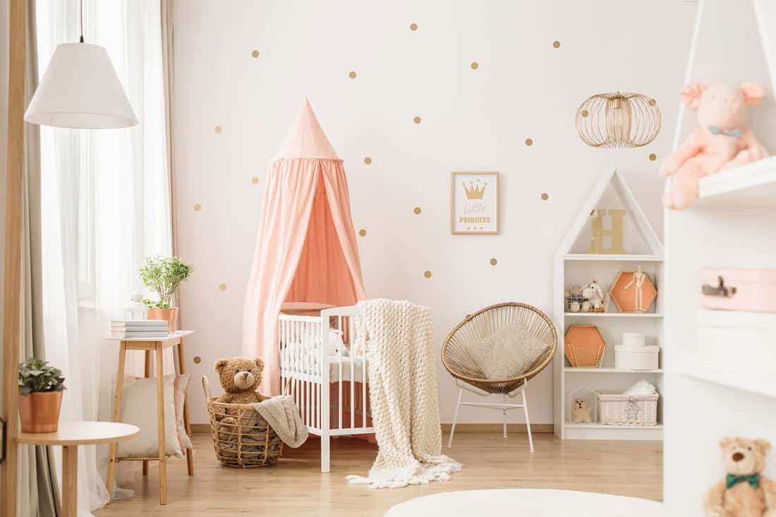 Canopied cradle between gold armchair and basket with teddy bear in pink baby's bedroom interior
