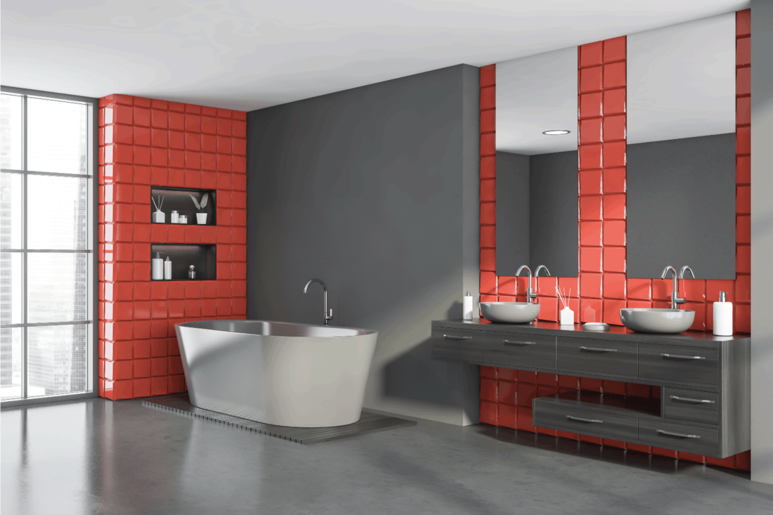 Corner of stylish bathroom with gray and red tile walls, comfortable double sink standing on dark wooden countertop with two mirrors and bathtub