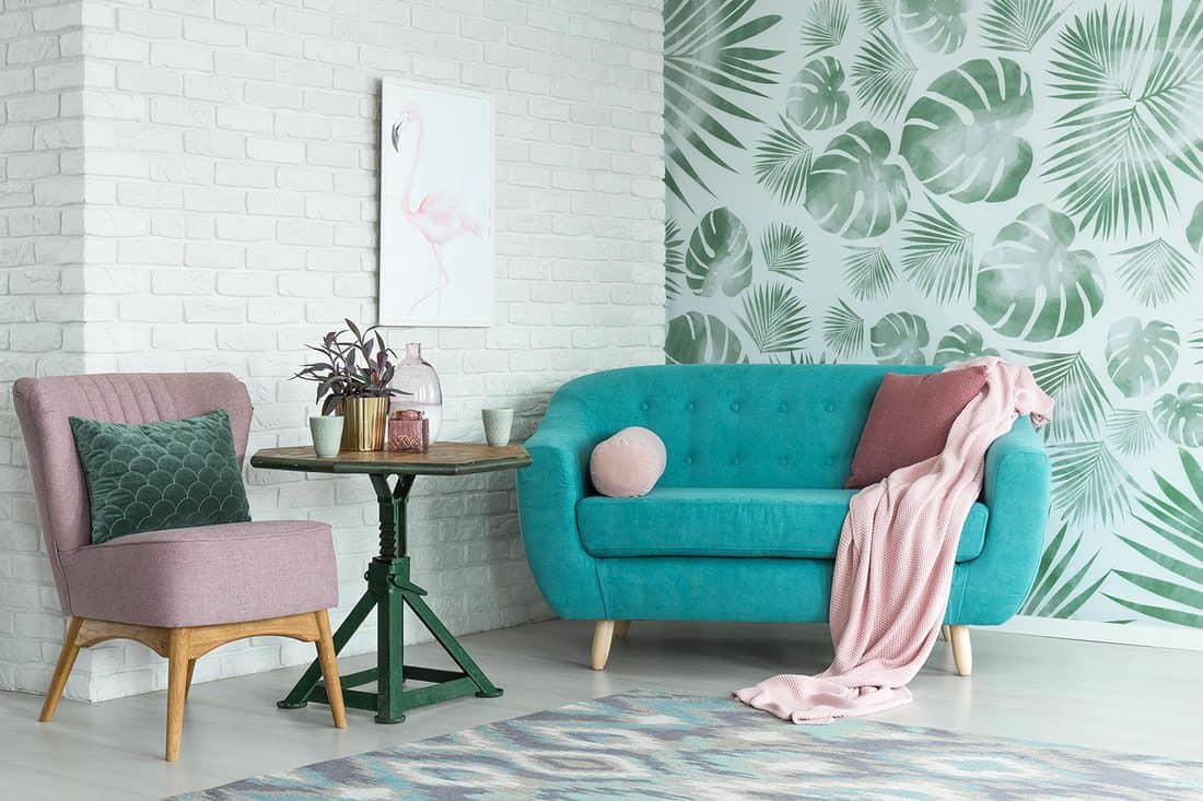Green table with plant between pink chair and blue sofa in floral living room with wallpaper and poster