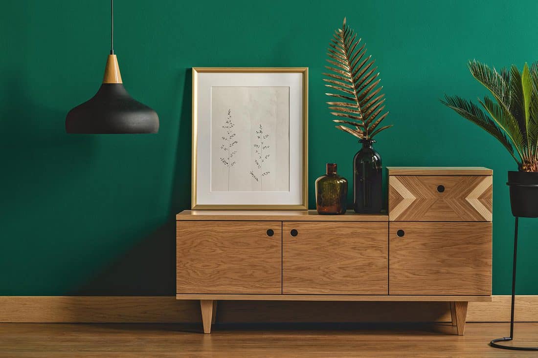 Industrial pendant light next to a stylish dresser and an art poster in a golden frame by a dark green wall of a modern bedroom