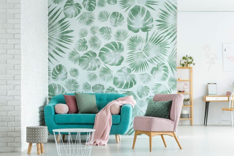 Pink armchair and blue settee with blanket in bright living room with stool and white table against leaves wallpaper, Does Wallpaper Need To Acclimate?