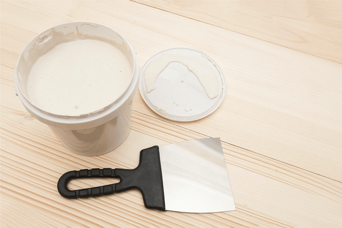 Spatula and a bucket of white putty on wooden boards
