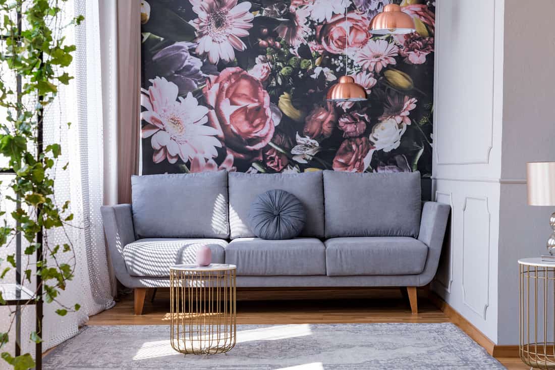 Sunlit, gray sofa by a floral print wall in the nook of a feminine living room interior with golden accessories, Does Wallpaper Make A Room Look Bigger Or Smaller?