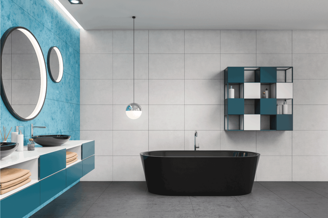Side view of stylish bathroom with white and blue tiled walls, tiled floor, comfortable double sink standing on blue countertop with round mirrors and black bathtub