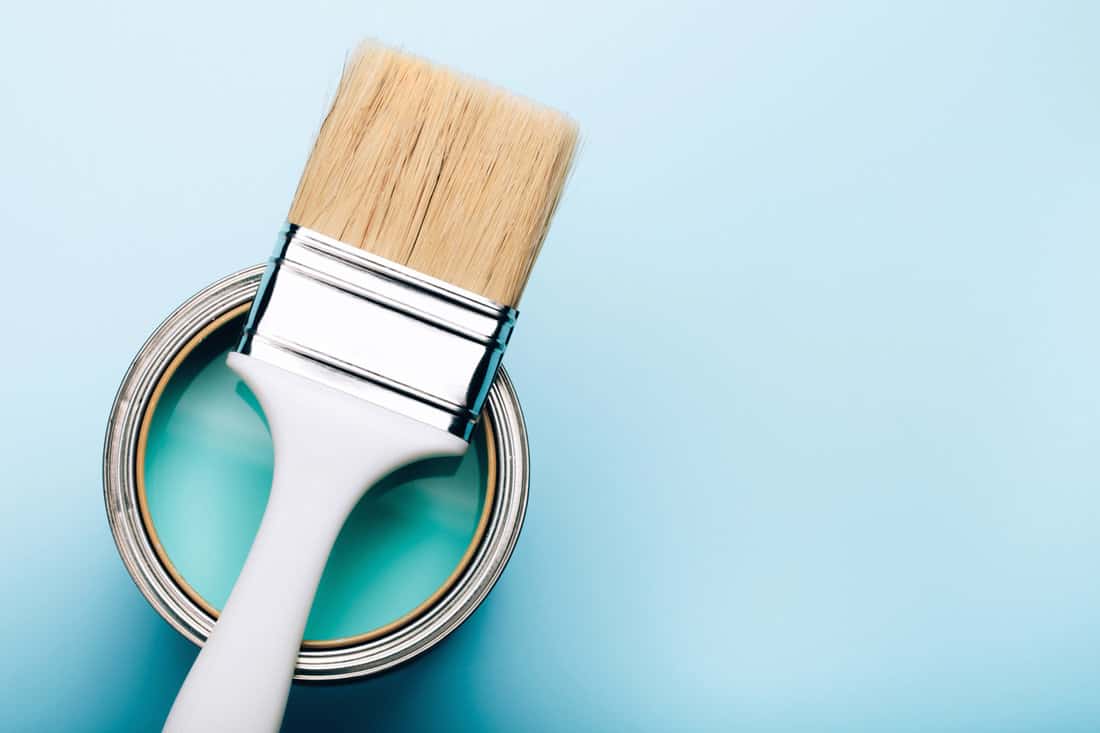Brush with white handle on open can of turquoise paint on blue pastel background. Renovation concept.