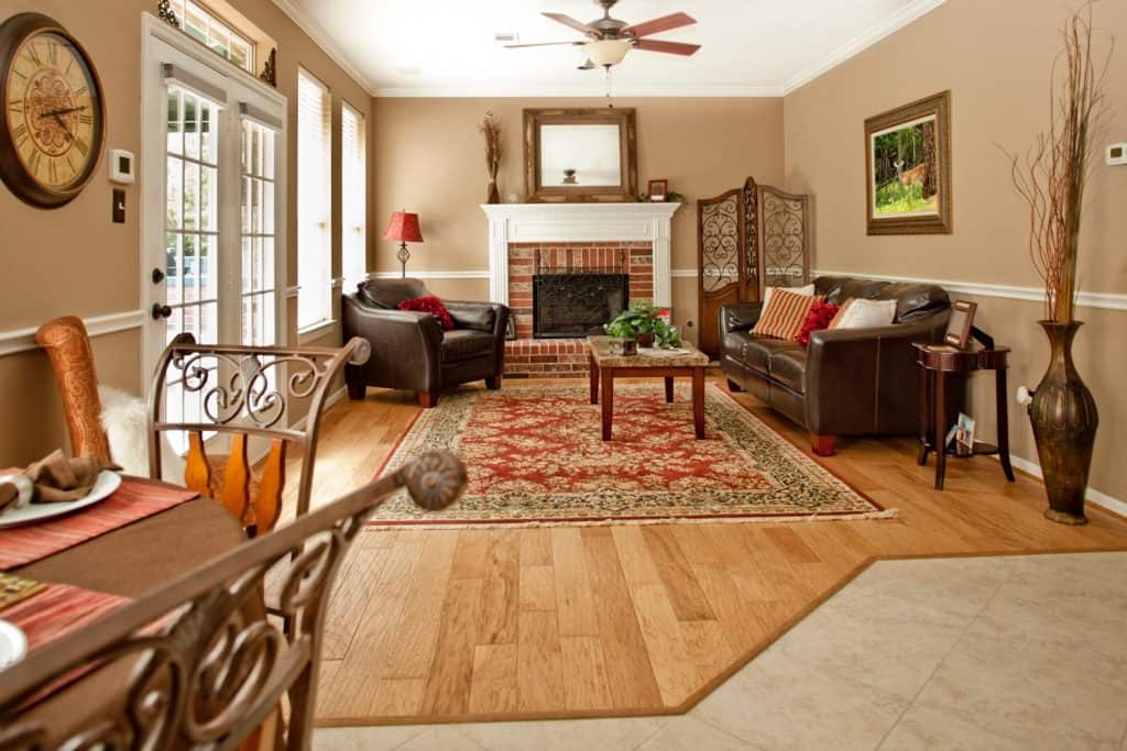 Lovely living room area of showcase home. Decor is red and tan color scheme. Seating area, fireplace. Breakfast table in foreground. Hardwood, tile floors.