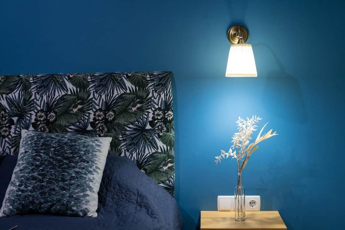 Sconces lamp on blue wall and flowers at nightstand table near comfortable bed, How High Do You Put Bedside Sconces?