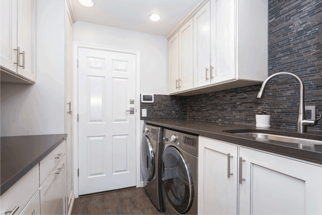 White laundry room boasts white shaker cabinets, brown grey glass tiled backsplash and modern washer and dryer placed under brown counter.