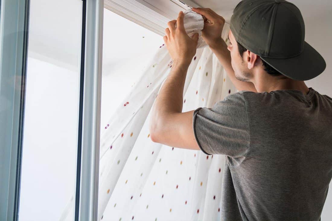 Young man installing curtains over window, How High Should You Hang Curtains? [By Ceiling Height]