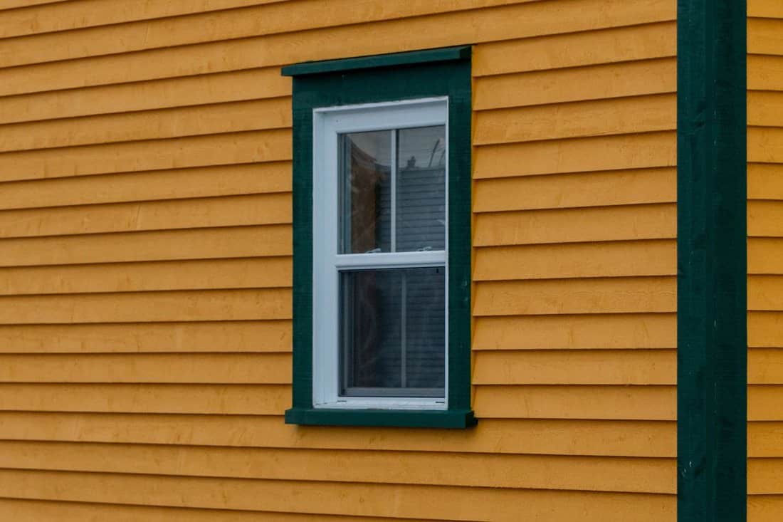 The exterior corner wall of a vibrant yellow colored wood clapboard siding. There's a single double hung window with green and white trim in the house. The corner of the building is thick green board.