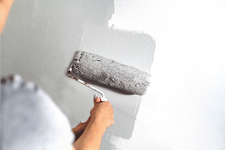 Human hand with large roller of paint begins to paint a white wall. Should You Paint The Ceiling Or Walls First