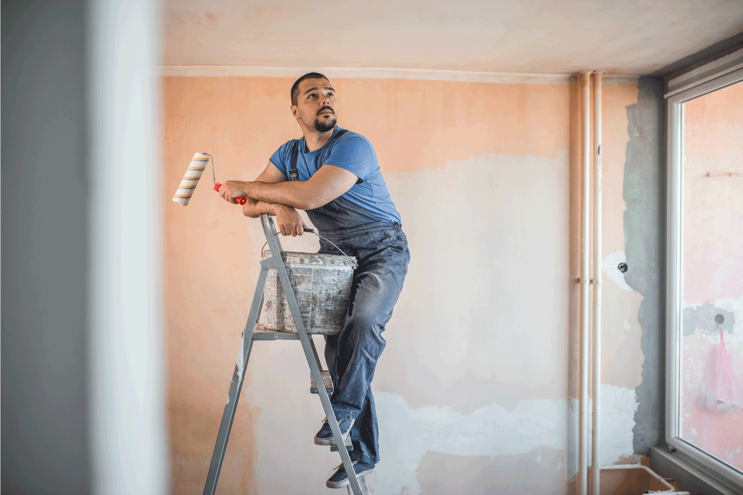 painter standing in room in coveralls holding paint roller and bucket