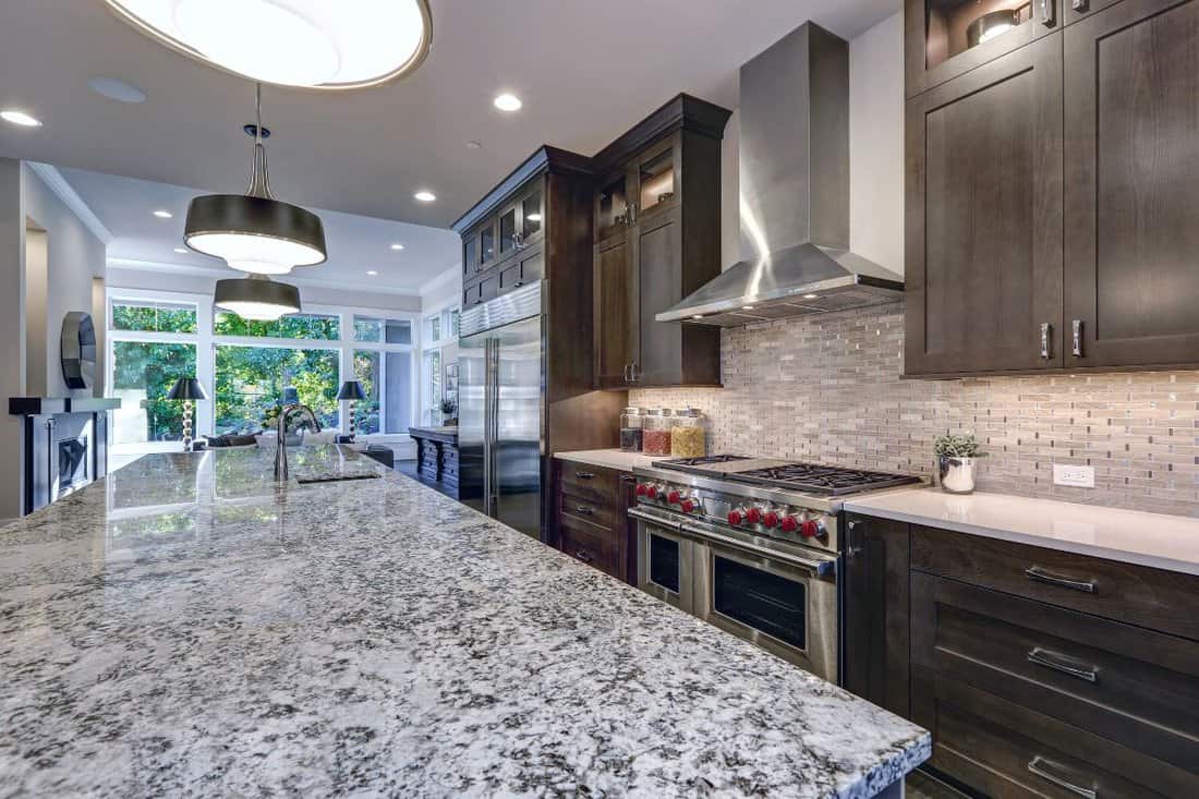 Modern kitchen with brown kitchen cabinets, oversized kitchen island, granite countertops, stainless steel hood over six burner Range and beige backsplash