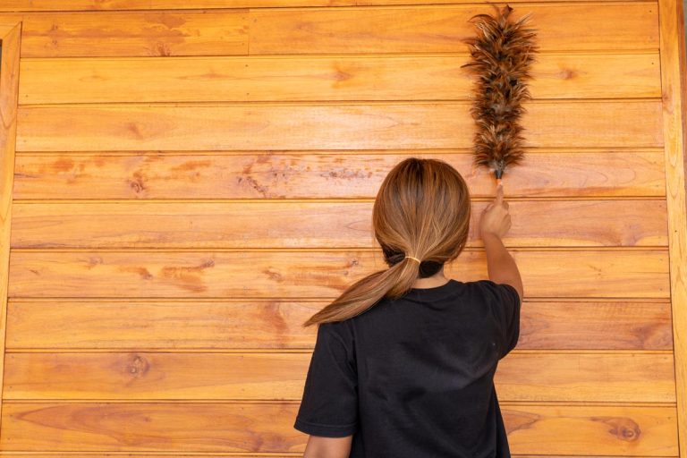 A woman dusting a wood wall with a feather duster, How To Clean Cedar Wood Walls [Inc. Mildew]