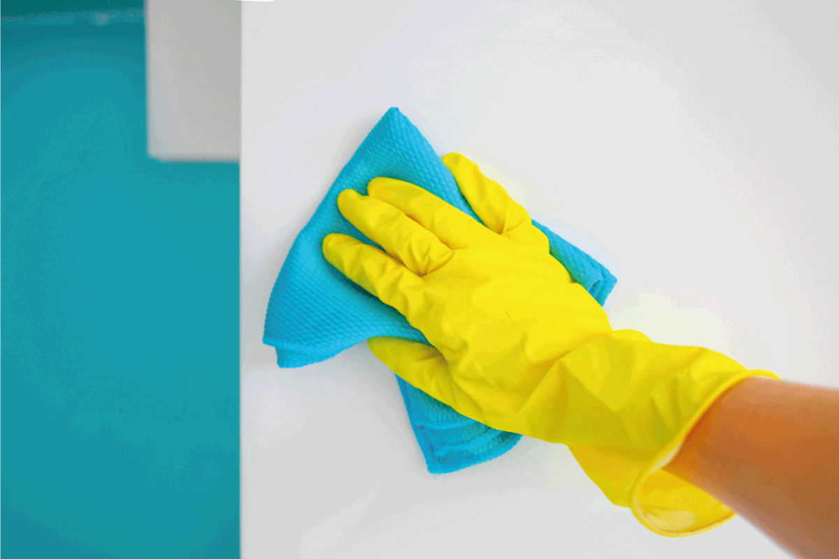 A female hand in yellow rubber gloves wipes a modern white wall with a microfiber cloth. How To Clean Dust Off Walls After Floor Sanding [4 Easy Methods!]