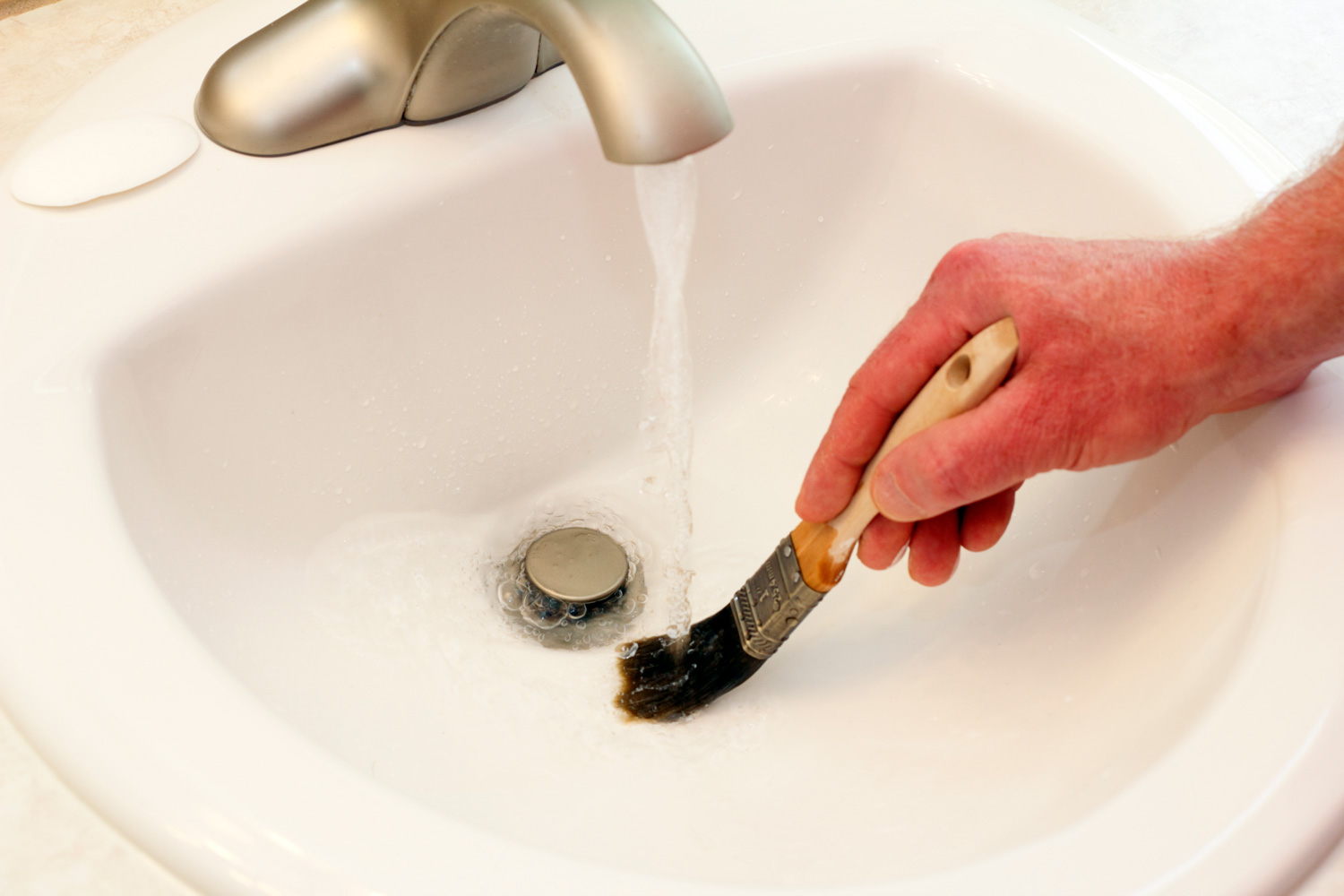Male caucasian hand rinsing a one inch wooden handle paintbrush under running water in a white bathroom sink.