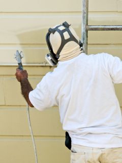 A man on a ladder uses spray gun to paint the exterior of a wooden building, What's The Best Finish For Exterior Paint