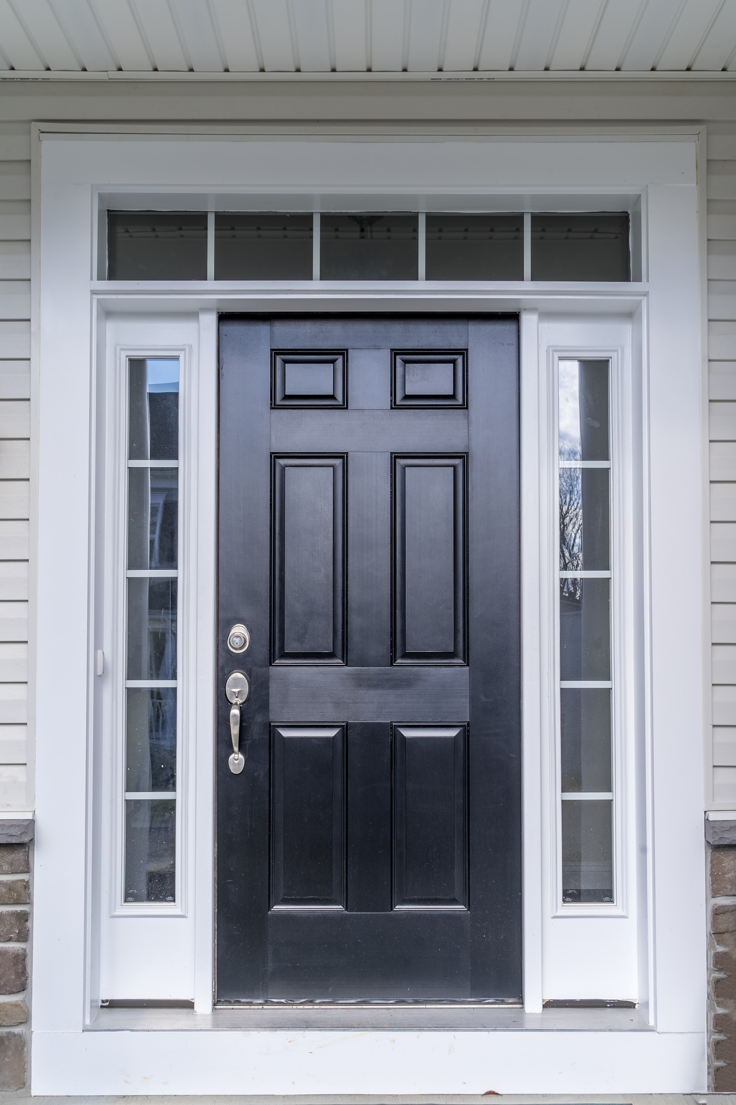 Classic fiberglass prehung black front door, raised panels, white frame, sill, jamb separating the divided sidelights, transom windows separated by narrow white grilles