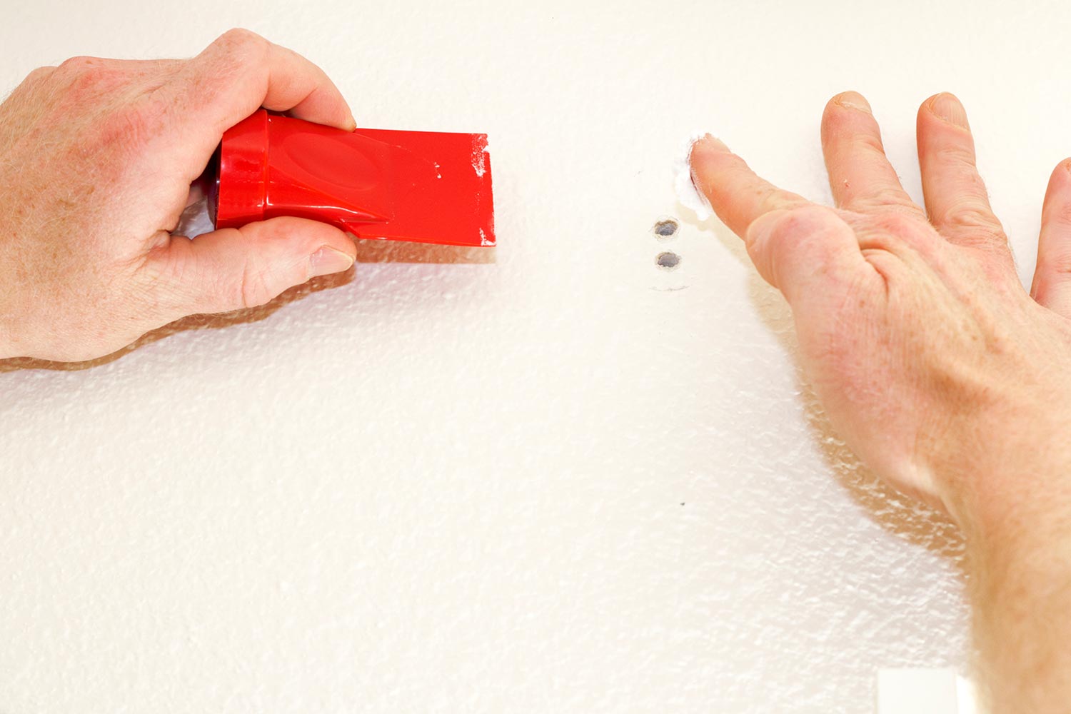Hand about to apply white spackling paste to two holes in the wall while holding on other hand a red tool to smooth spackling