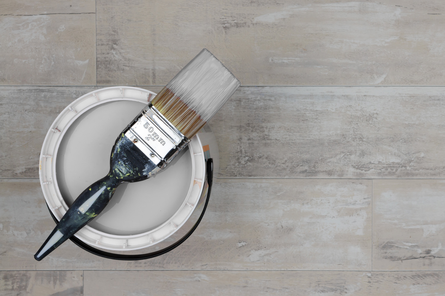Looking down on loaded paintbrush placed across an open can of grey paint stood on a shabby style wood floor