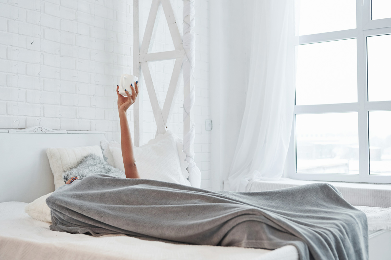 Woman lying under blanket and raising hand up with white cup of coffee in it.