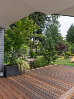 A perspective view of a contemporary Pacific Northwest home with a deck bridging a pond that leads to a pair of modern yellow loungers in a landscaped yard, Can You Sand A Deck After It Rains?