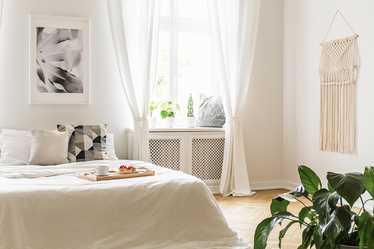 Breakfast tray with pastries and coffee on a cozy, white bed in a bright and peaceful bedroom interior with plants on a window sill seat