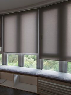 Motorized roller shades in the interior. Automatic roller blinds beige color on big glass windows. Home luxury curtaines are above the windosill with pillows. Summer. Green trees outside. - Should Blinds Match Throughout House