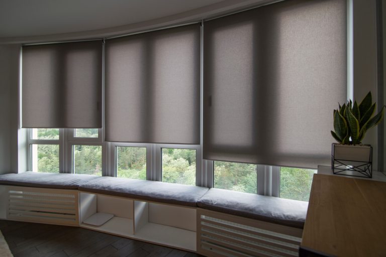 Motorized roller shades in the interior. Automatic roller blinds beige color on big glass windows. Home luxury curtaines are above the windosill with pillows. Summer. Green trees outside. - Should Blinds Match Throughout House