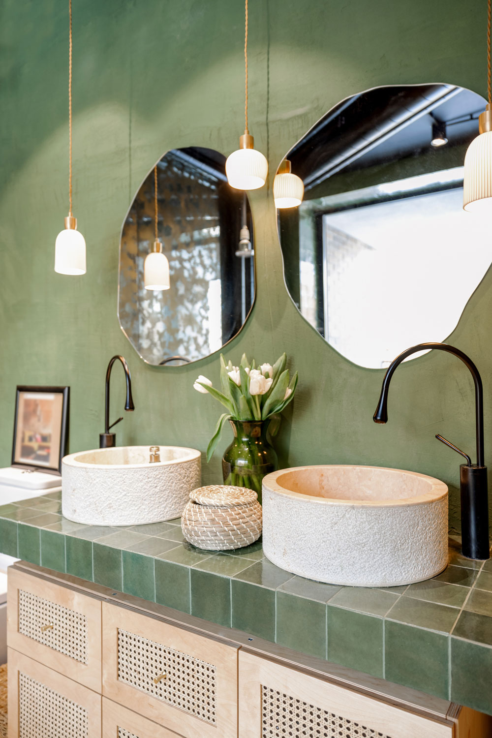 Matte green painted bathroom walls with dangling lamps and a green tiled vanity with irregular shaped mirrors