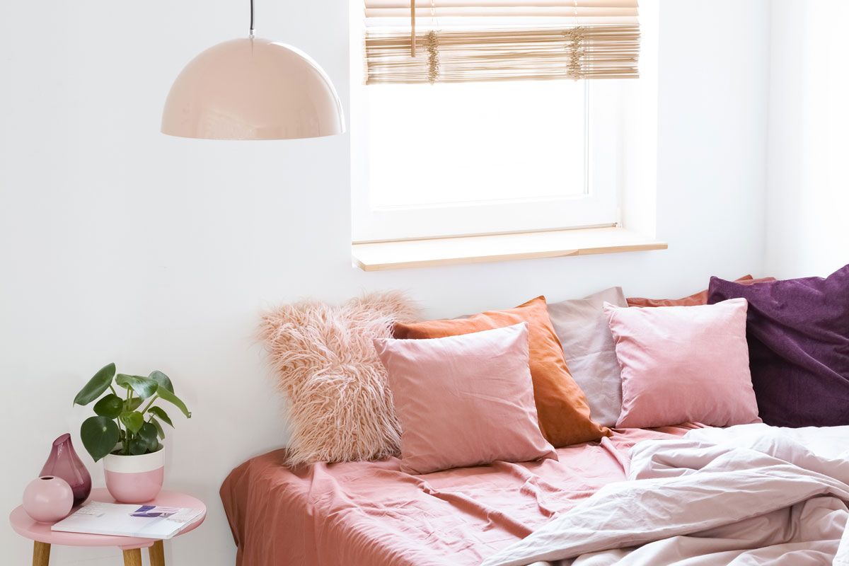 Pink pillows on bed next to table with plant in bright bedroom interior with window and lamp