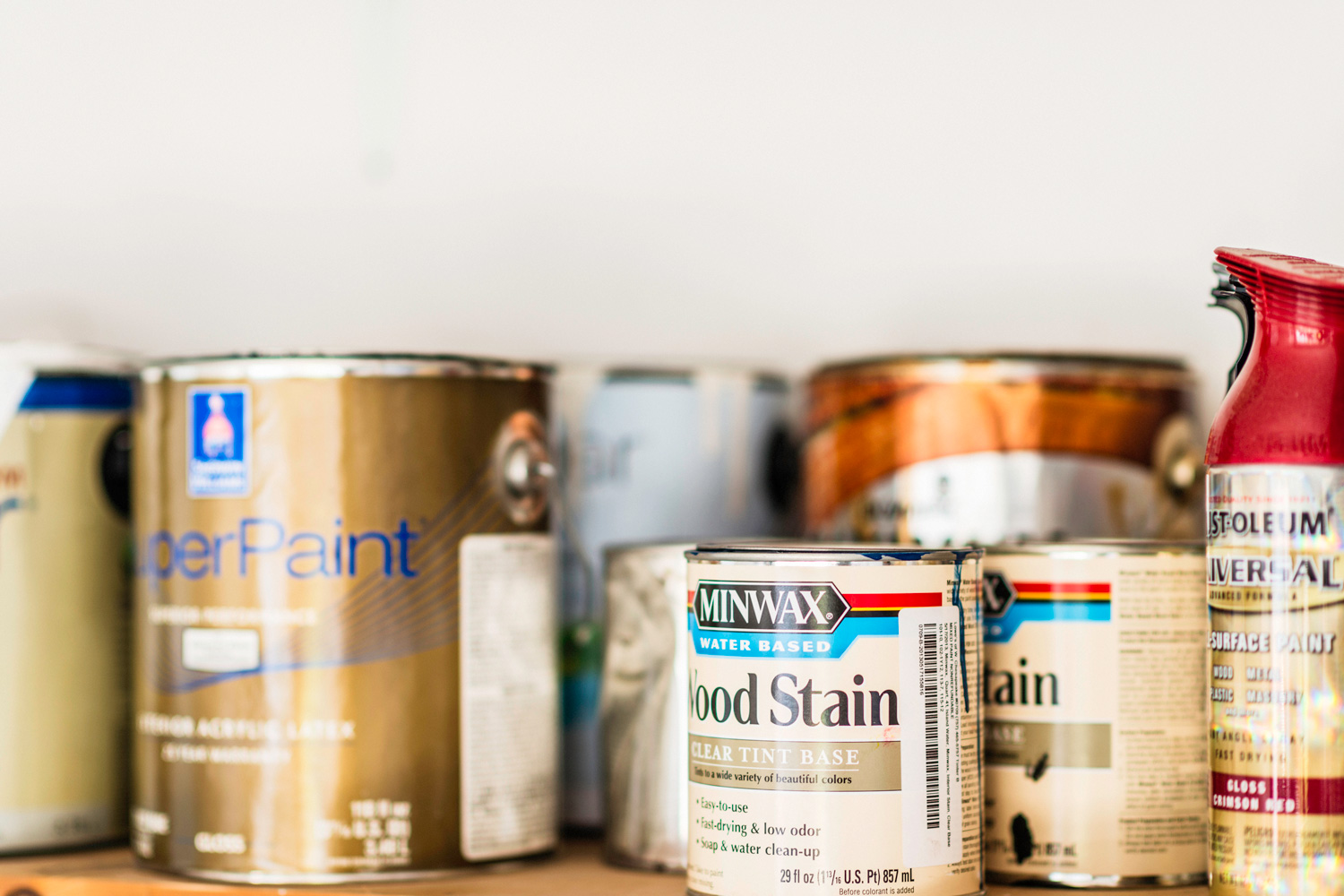 A horizontal shot of a collection of opened and unopened American brand cans of paint, wood stain and paint sprays organized neatly on a wooden shelf. The brands include cans of indoor house paint by Sherwin Williams, spray paint by Rustoleum, and wood stain by Minwax.