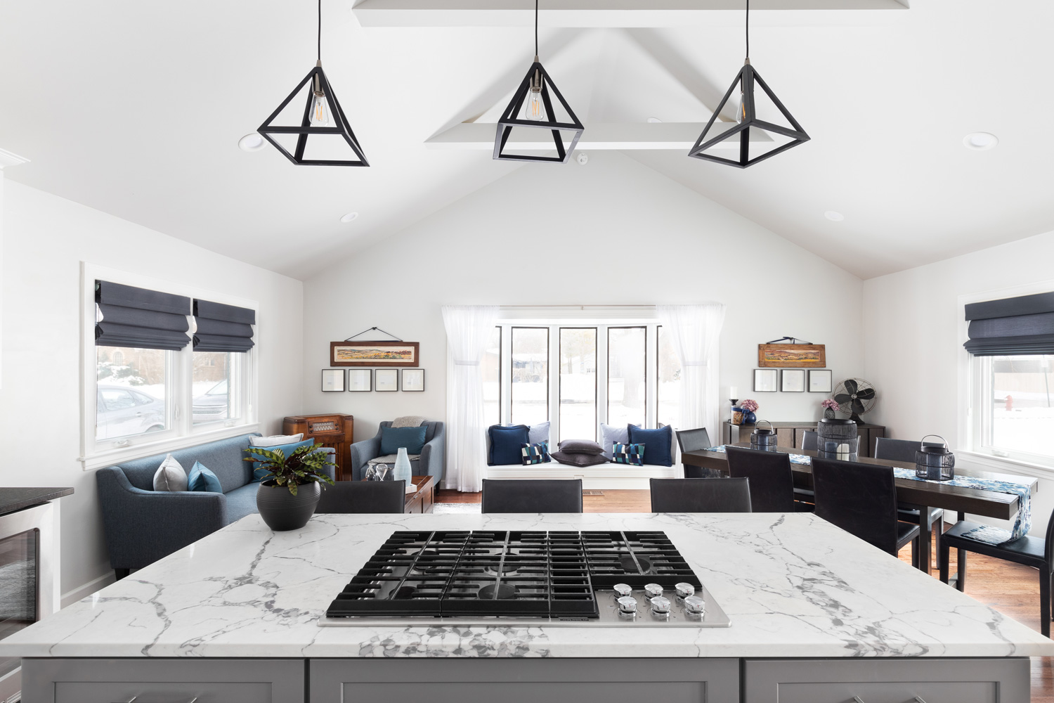 A luxury kitchen with lights hanging above the island looking towards a modern farmhouse living and dining room. White beams hang from the cathedral ceiling.