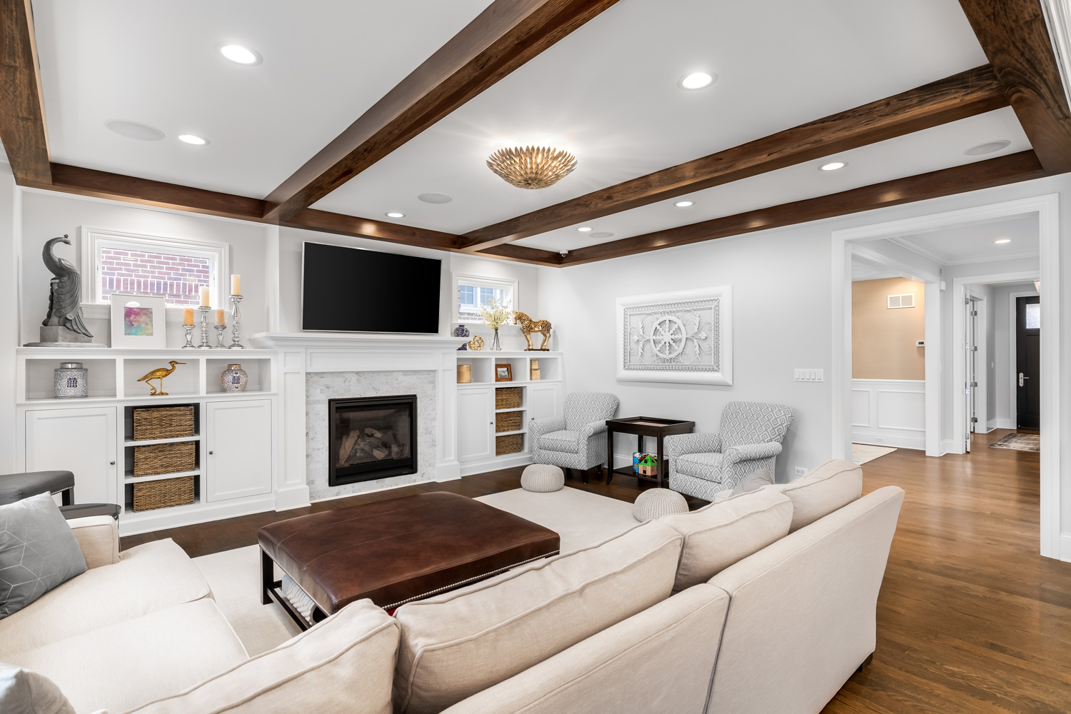  A luxury, modern farmhouse living room with wood beams on the ceiling, a tiled fireplace surrounded by built in shelves and furniture on the hardwood flooring.