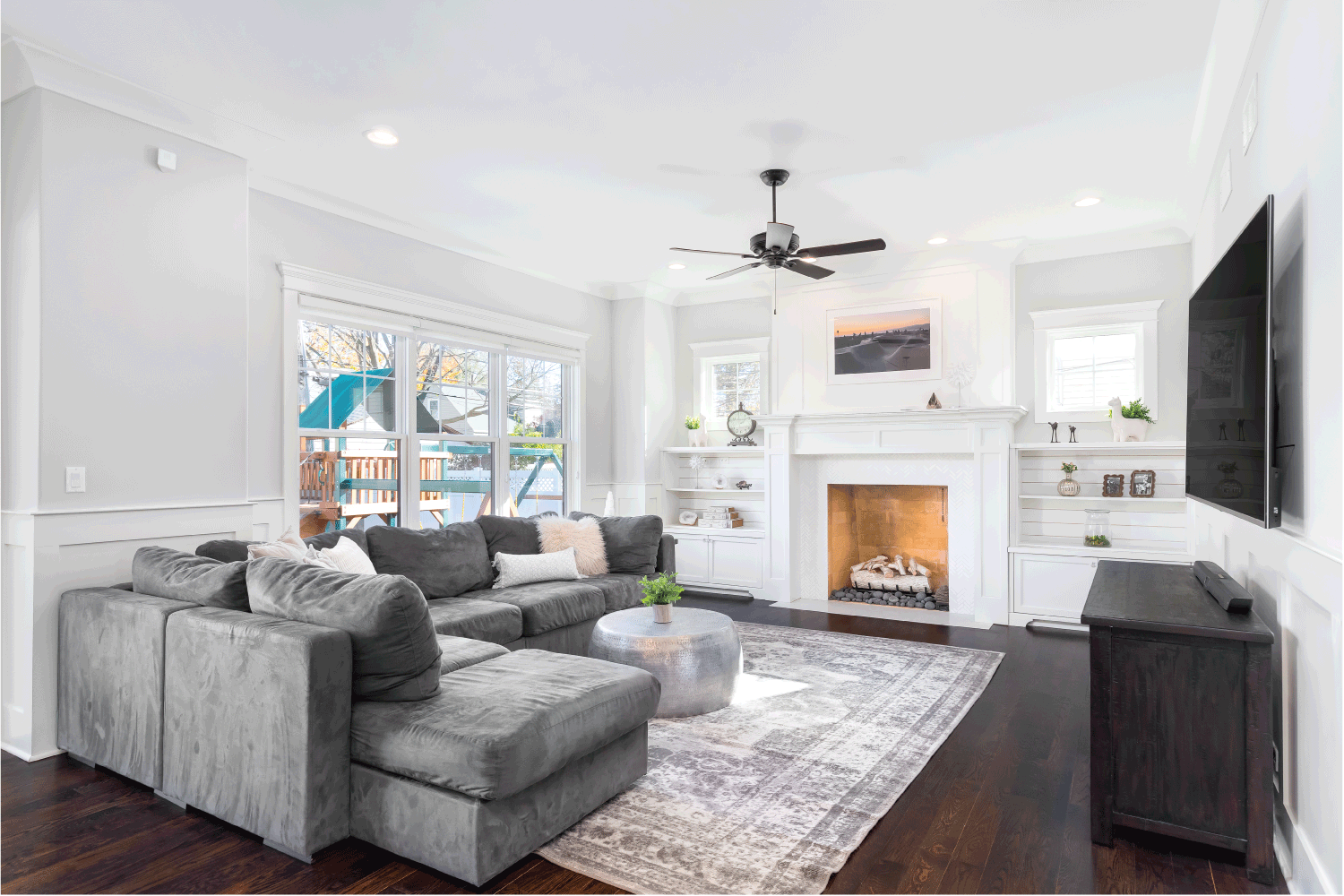 A cozy, white living room with a large grey sectional couch, an area rug on hardwood floors, and a white fireplace with built-in shelves.