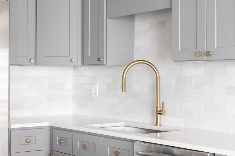 A kitchen sink detail shot with a gold faucet, marble backsplash, grey cabinets, and gold hardware, What Backsplash Goes With Taj Mahal Quartzite?