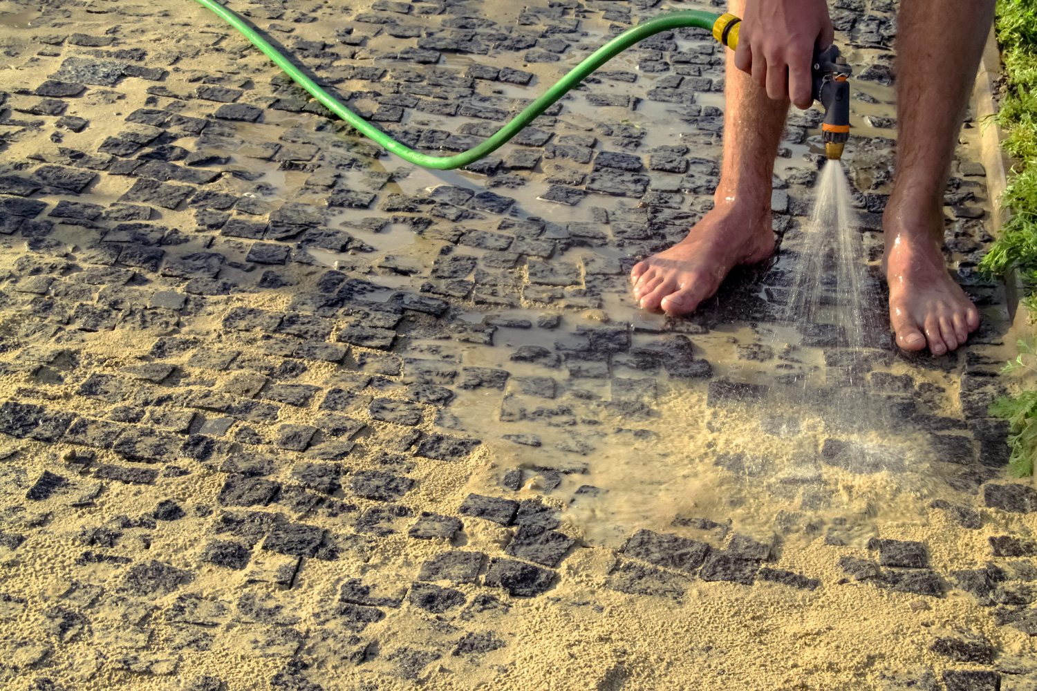 A man washes his bare feet from a watering hose, standing on a sandy paving stone, in the summer outdoors 