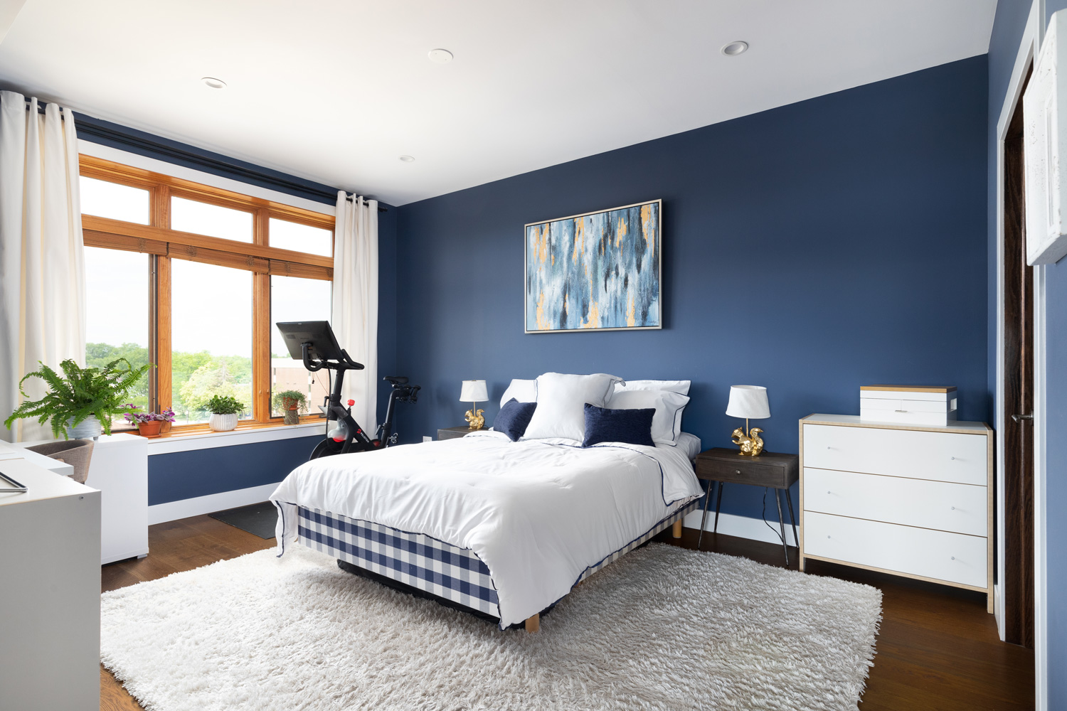A navy blue bedroom with wood framed windows a bed sitting on a rug and hardwood floors, and a Peloton.