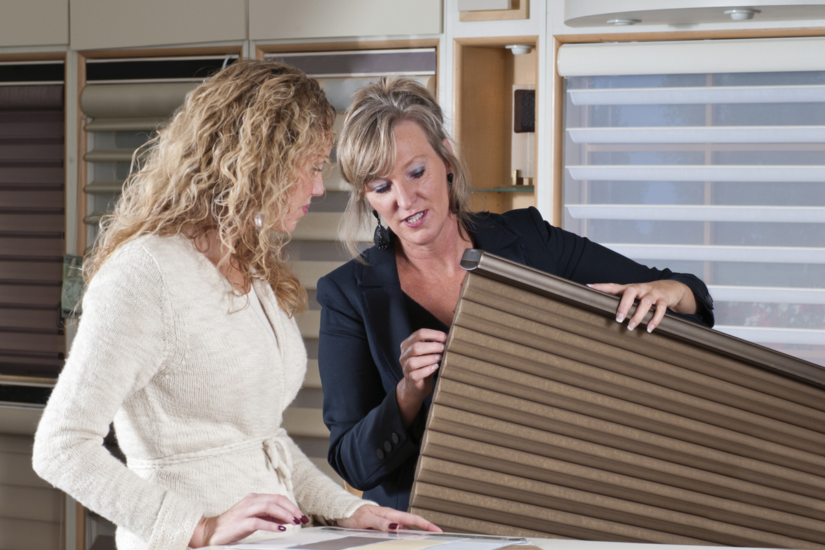 A saleswoman at a home furnishings store assists a female shopper with designer window blinds.