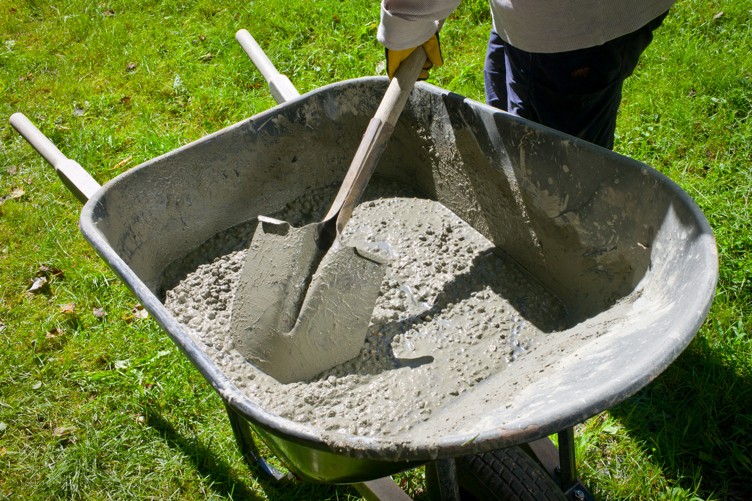 A woman uses a shovel to mix cement by hand in a wheelbarrow on a bright, sunny morning.